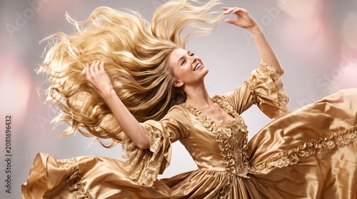 Joyful young woman in golden baroque satin dress with long flowing blonde hair tossed dramatically mid-air, smiling against soft studio background, perfect for hair care beauty advertising
