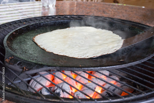 Outdoor Pancake Frying on a Charcoal Barbecue Grill. Fresh pancake browning evenly on a cast iron pan above glowing coals during outdoor cooking.