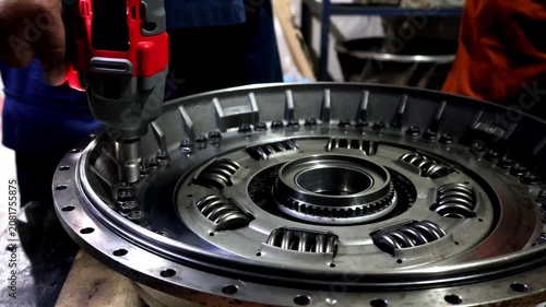 Close up of a technician's hand operating a cordless impact driver to tighten bolts on a large metallic clutch assembly or torque converter.