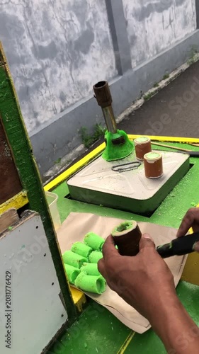 Street vendor preparing traditional green cylindrical snack called 'Kue Putu' on a small portable gas stove.
