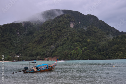 A traditional Thai long-tail boat floats on emerald waters beneath mist-covered limestone cliffs near Phi Phi Don Island in Krabi Province after tropical rainfall