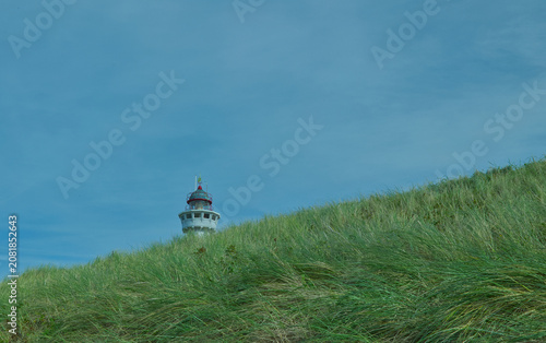 Spitze eines weißen Leuchtturms hinter einer grünen, grasbewachsenen Düne vor blauem Himmel