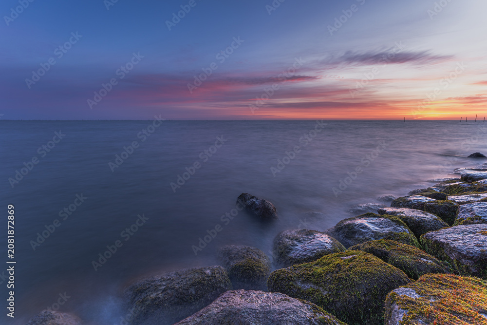 Naklejka premium Markermeer, Volendam, Netherlands. Long exposure technique creates a smooth, silky appearance on the water surface, while the moss-covered rocks in the foreground add texture, depth and visual interes