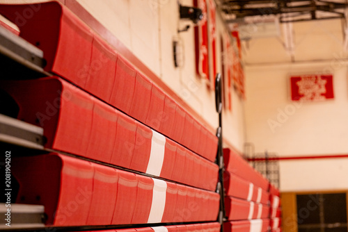 Quadro em tela Stacked red folded bleacher seating panels rest against a tan wall in a school gymnasium, showing the compact, retracted state of a multi purpose sports facility