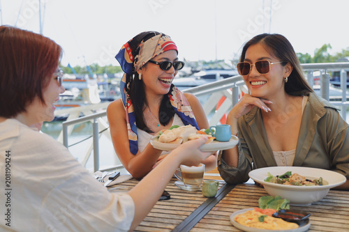 Three cheerful young women enjoying lunch together at a marina restaurant, smiling and sharing food during a casual weekend outing