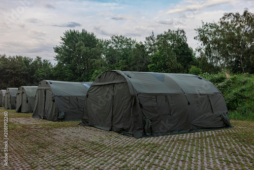 Olive military tents line a green field under a cloudy sky, an army camp during a field exercise.