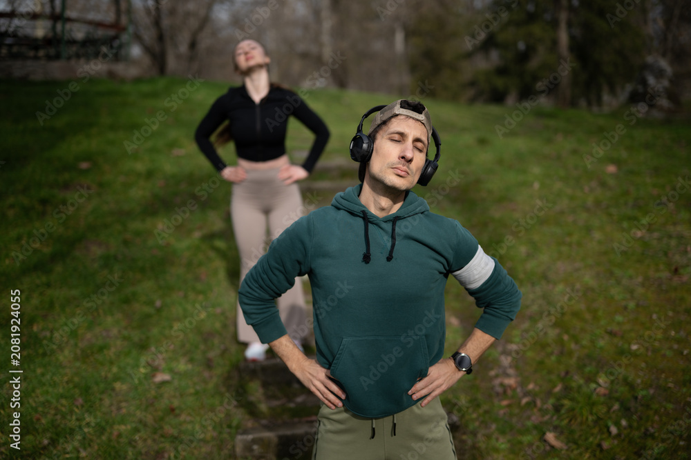 Naklejka premium Man wearing headphones stands with hands on hips and eyes closed in a grassy park. A woman in the background stretches with hands on hips on a step pathway.