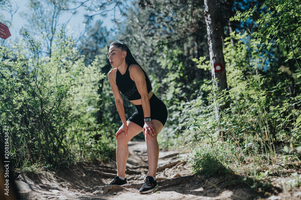 Obraz premium A young woman wearing athletic gear catches her breath while leaning on her knees on a forest trail. She pauses during a trail run through sunlit woods to rest and recover.