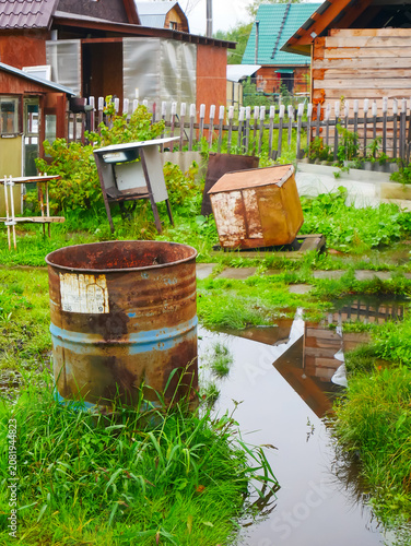 Rusty metal barrel standing in a flooded garden backyard. Discarded scrap and old trash scattered on grass after heavy rain. Environmental pollution and neglected village property.