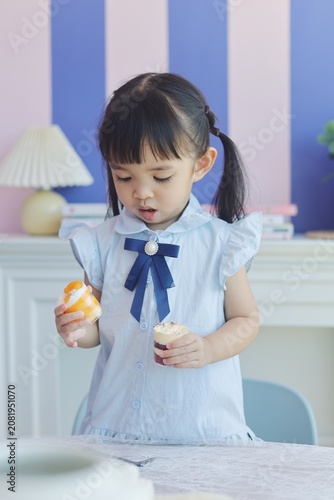 little Asian girl enjoying colorful mini cakes at the table, happily eating dessert with a spoon. 