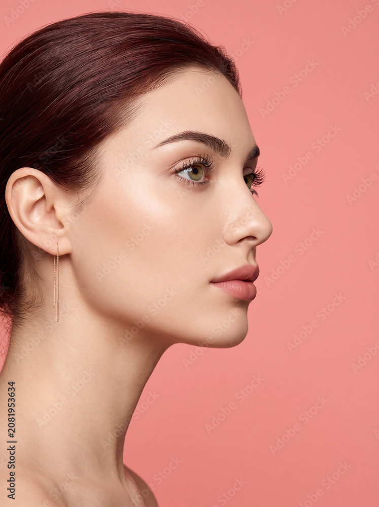 Fototapeta premium Elegant studio portrait of a young woman in side profile featuring flawless glowing skin, soft natural makeup, and a warm pink background with premium beauty styling.