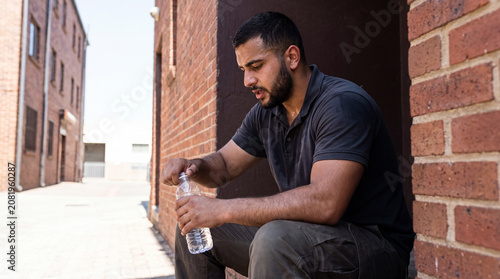 Exhausted worker opening a water bottle in an urban alleyway, sweating and trying to rehydrate during a hot day