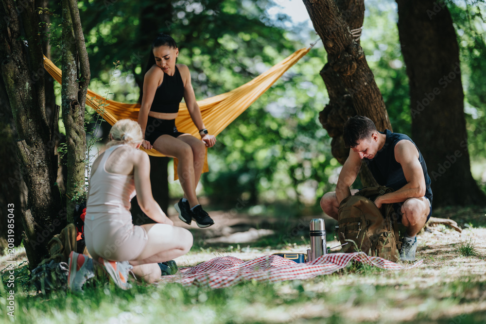 Naklejka premium Three young friends rest during a hiking break in a shaded forest clearing. They sit on a picnic blanket and a hammock while checking gear and enjoying nature.