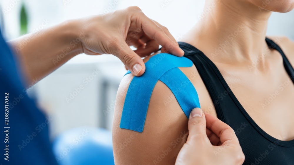 Obraz premium A detailed shot captures a professional applying vibrant blue kinesiology tape to a woman's shoulder, aiming to alleviate muscle discomfort and support recovery