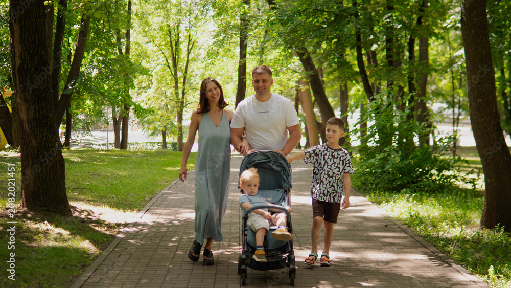 Naklejka premium Father and mother stroll along tree-lined paved park path with toddler seated in stroller and older child walking beside them; dappled sunlight filters