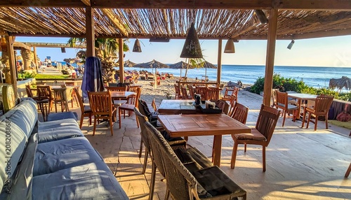 A serene beachside patio with wooden tables and chairs under a thatched roof