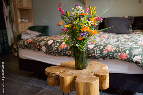 Bright mixed wildflower bouquet in clear glass vase on carved wood table with patterned bedspread . High quality photo