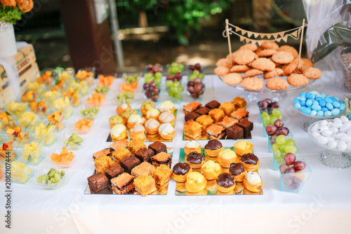 A bright dessert table featuring mini pastries, fresh fruits, and colorful candies. The setting suggests a festive outdoor event in natural light.