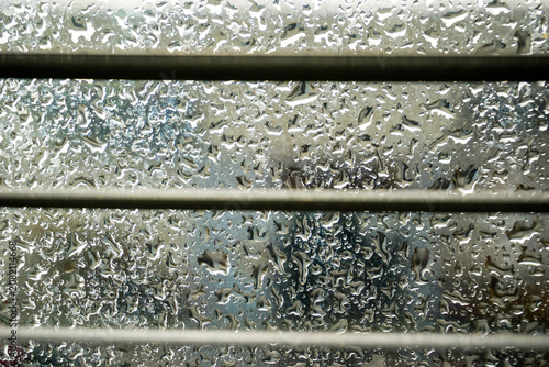 Close-up view of raindrops on a glass window with metallic blinds. The blurred background gives a dreamy and moody atmosphere on a rainy day.
