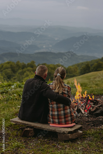 Couple sitting by a campfire overlooking misty mountain landscape at sunset. Romantic outdoor camping scene with scenic views, perfect for travel, adventure, hiking, nature and lifestyle concepts.