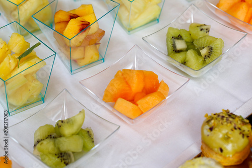 Colorful assortment of diced fruits including kiwi, melon, and pineapple served in small transparent containers on a white tablecloth.