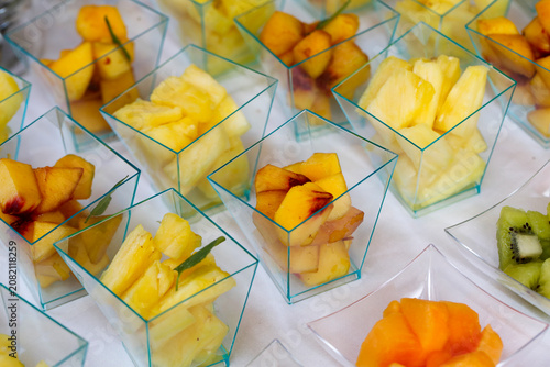 Colorful diced fruits including pineapple, peach, kiwi, and melon arranged neatly in small plastic cups on a bright white tablecloth.