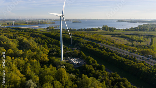 Aerial view of a wind turbine standing amidst lush green trees near a highway and the coastal waters in Bruinisse, Zeeland, Netherlands.