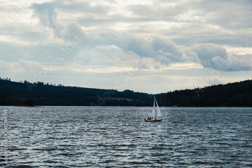 Small sailboat sailing on a wide lake under cloudy sky with forested hills in the background, concept of outdoor recreation, travel, solitude and calm lakeside nature.