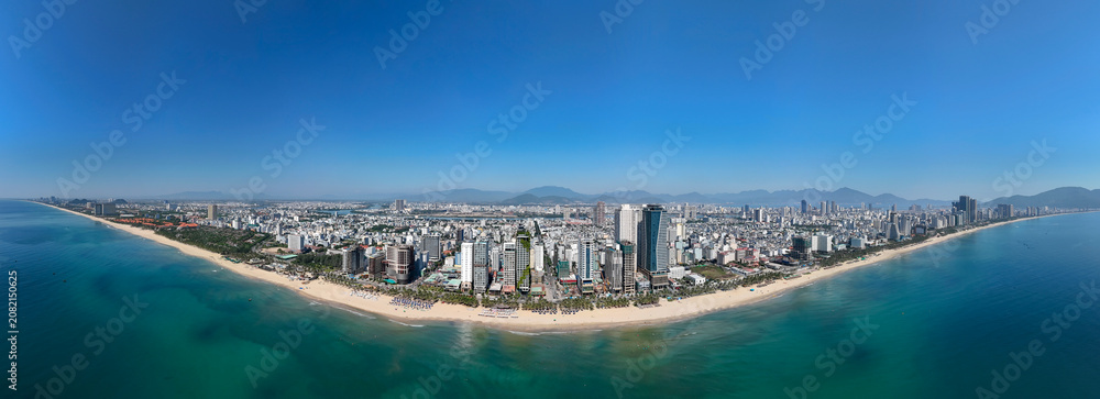 Fototapeta premium Expansive aerial view of Da Nang city buildings lining the vast My Khe Beach with turquoise ocean under a bright blue sky. Mountains rise in the distance