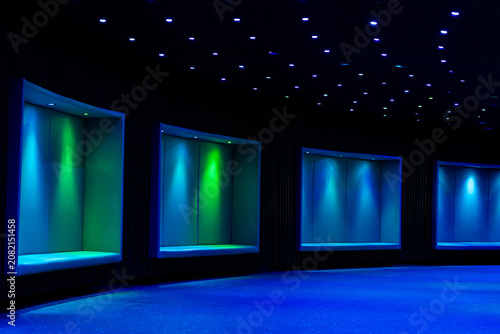 Dark modern interior with blue and green neon lighting, built in seating areas and small ceiling lights. Atmospheric architectural scene with futuristic design, soft glow and empty indoor space.