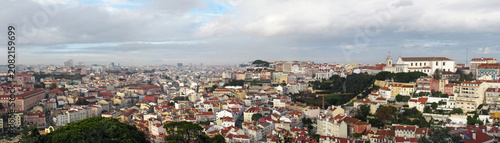 Wide panoramic aerial view of Lisbon cityscape with historic white buildings and terracotta roofs under a cloudy dramatic sky.        