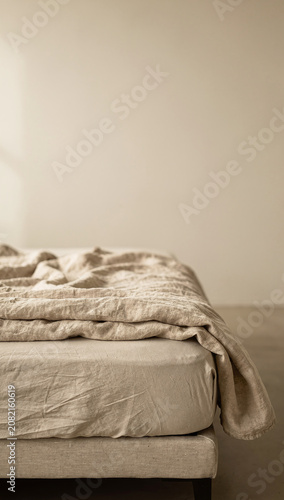 Unmade bed with crumpled beige linen sheets and blanket in a minimalist bedroom