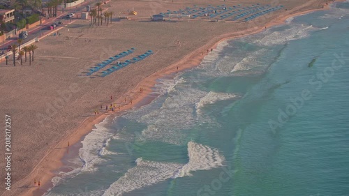 Footprints line the shore as people walk along the beach. Sun loungers are set up on the sand. Waves crash gently on the beach. The sun shines brightly on the water.