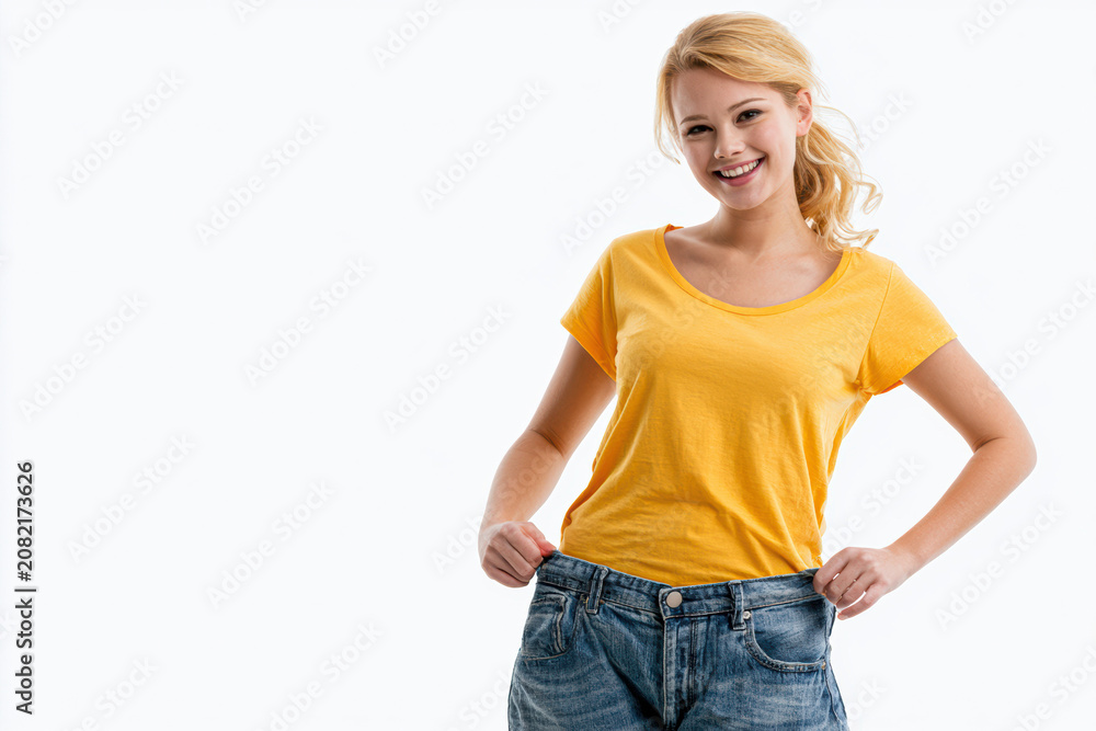 Fototapeta premium Happy young woman in loose jeans and yellow t-shirt showing weight loss with a smile on a white background.