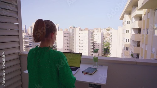 A person stands on a balcony looking out at the city. They are working on a computer while enjoying the view. The sun is shining and it is daytime.