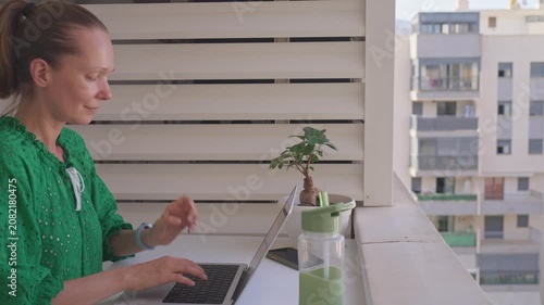 A woman sits on a balcony with a laptop. She types and looks at the screen. A small plant is beside her. The sun shines as she focuses on her work in the afternoon.