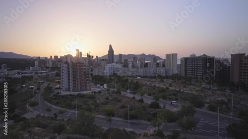 The sun rises over a city skyline, casting light on the buildings and mountains. People start their day as shadows fade. Calm morning signals a new beginning.