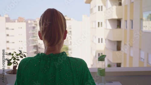 A person is sitting at a table on a balcony. They are focused on their laptop while looking out at the city. Sunlight shines during the day, highlighting the buildings around them.