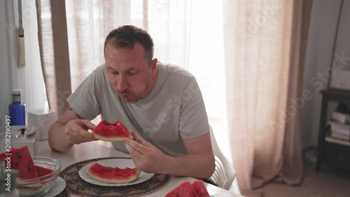 A person sits at a table in a home. They are enjoying slices of watermelon. Sunlight shines through the window. The table has a plate of watermelon pieces.