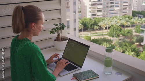 A person sits at a table on a balcony and types on a laptop. The sun shines on the city while the person smiles and enjoys the view. Green plants add to the setting.