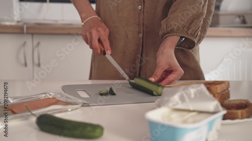 A person is chopping cucumber on a cutting board in a kitchen. The person focuses on the task while holding a knife. The atmosphere shows ingredients prepared for a meal.