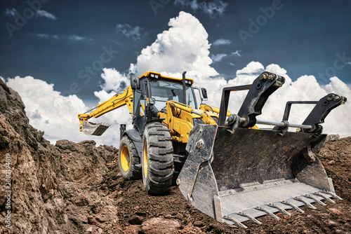 An excavator loader is working at a construction site. It is clearing dirt and rocks while clouds fill the sky. Heavy equipment is in use for a project