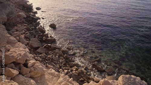 The camera shows a rocky shoreline by the sea as the sun sets. The water is calm and reflects the colors of the sky. Small waves wash against the rocks.