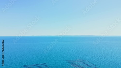 Mussel farm is located in the ocean with rows of long lines set in the water. Bright sky shows few clouds as sunlight reflects off the surface. Distant land can be seen.