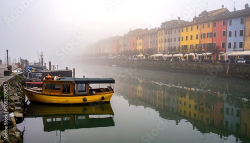 A serene harbor scene with a vibrant yellow boat on calm waters, lined by colorful buildings on a foggy day