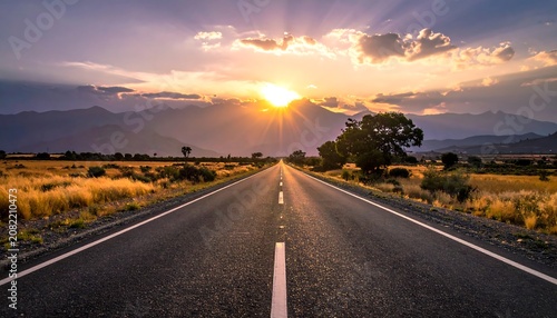 A serene highway stretches into the distance, lined with golden grass and trees on either side, under a vibrant sunset