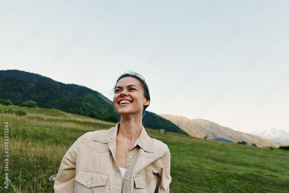 Fototapeta premium Smiling woman in casual jacket standing outdoors in a green field with mountains in the background. Concept of lifestyle, nature, happiness, and freedom in soft natural light.