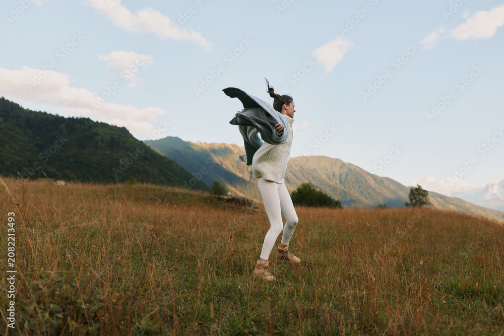 Fototapeta premium Young woman jumping in a grassy field with mountains in the background. Outdoor lifestyle concept with casual clothing and natural light during daytime.