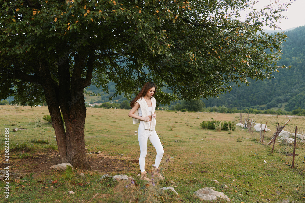 Fototapeta premium Woman standing beside a large tree in a rural meadow, wearing white clothing. Lifestyle portrait outdoors, peaceful countryside scene, natural light, relaxed pose, simple fashion.