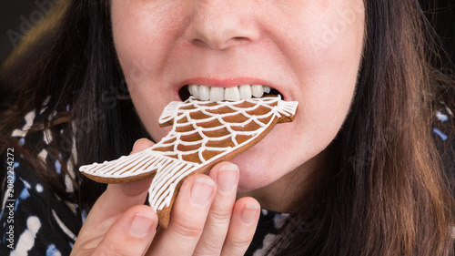 Closeup of a female face with gingerbread fish in mouth at eating Christmas pastry. Woman with long brown hair when is holding in hand and biting into sweet baked Xmas cookie decorated by white icing.